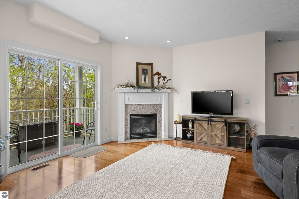 Cozy living room in a Traverse City condo featuring a gas fireplace, hardwood floors, large windows with views of surrounding woods, and a private balcony, ideal for relaxation.