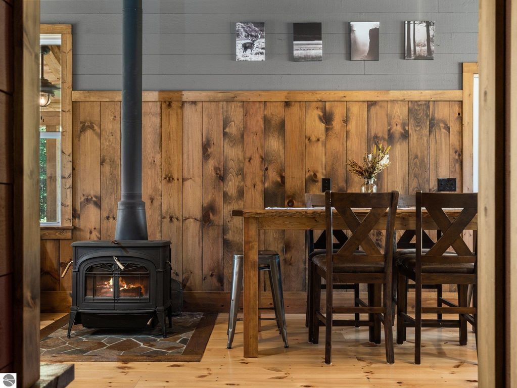 Cozy dining area with rustic wood paneling, a black woodstove, and a wooden table surrounded by X-back chairs, featuring decorative wall art and natural light from the window, reflecting the custom craftsmanship of the Lake City, MI home.