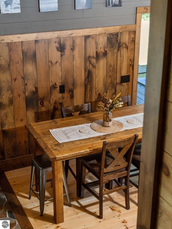 Rustic dining area featuring a wooden table with a white runner, surrounded by chairs and a vase of flowers, against a backdrop of wood-paneled walls, reflecting the custom craftsmanship of the Lake City, MI home.