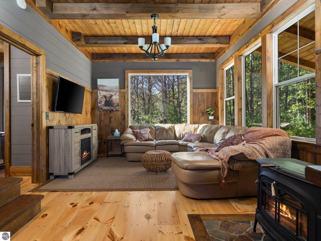 Cozy living room featuring a large sectional sofa, rustic wood accents, and a modern electric fireplace, with large windows showcasing views of the northern Michigan countryside.