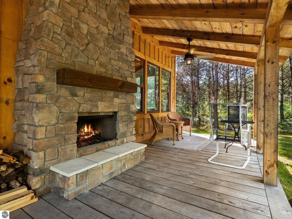Cozy covered porch with stone fireplace, wooden ceiling, and seating area overlooking serene northern Michigan landscape.