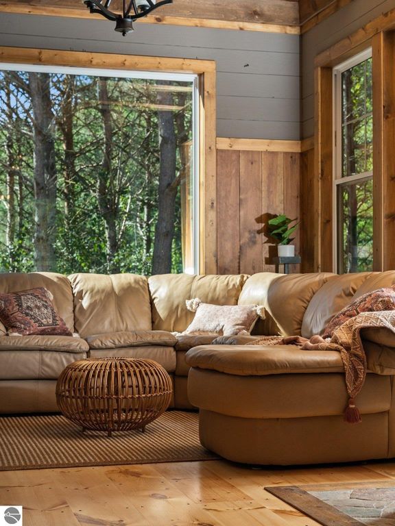 Cozy living area featuring a leather sectional sofa with decorative pillows, a round woven coffee table, and large windows showcasing the serene northern Michigan landscape.