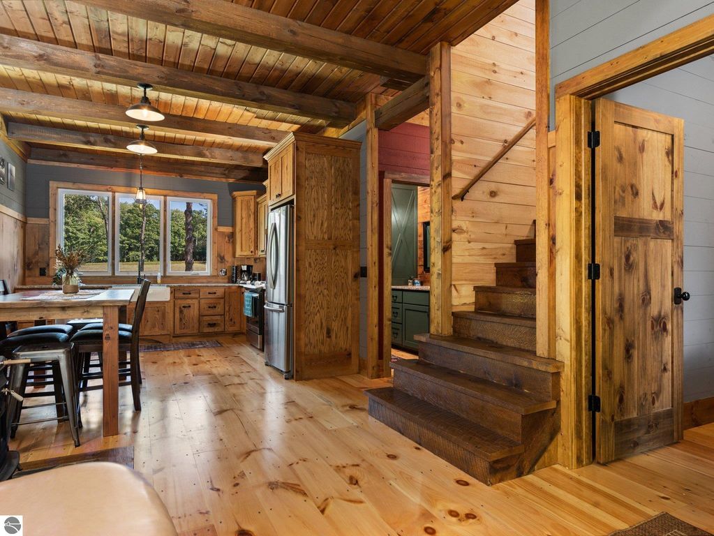 Cozy interior of a custom-built home featuring rustic wooden beams, handcrafted cabinetry, and an open-concept layout, showcasing a dining area and staircase, with large windows offering views of the surrounding nature.