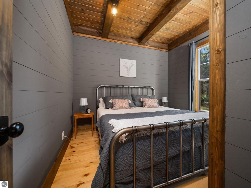 Cozy bedroom featuring a metal bed frame, gray walls, wooden ceiling beams, and natural light from a window, showcasing the custom craftsmanship of the home at 5800 W Moorestown Road, Lake City, MI.