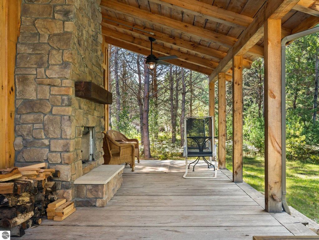 Covered porch with stone fireplace, wooden ceiling beams, and seating area surrounded by northern Michigan forest, showcasing outdoor living space of custom-built home on 10 acres.