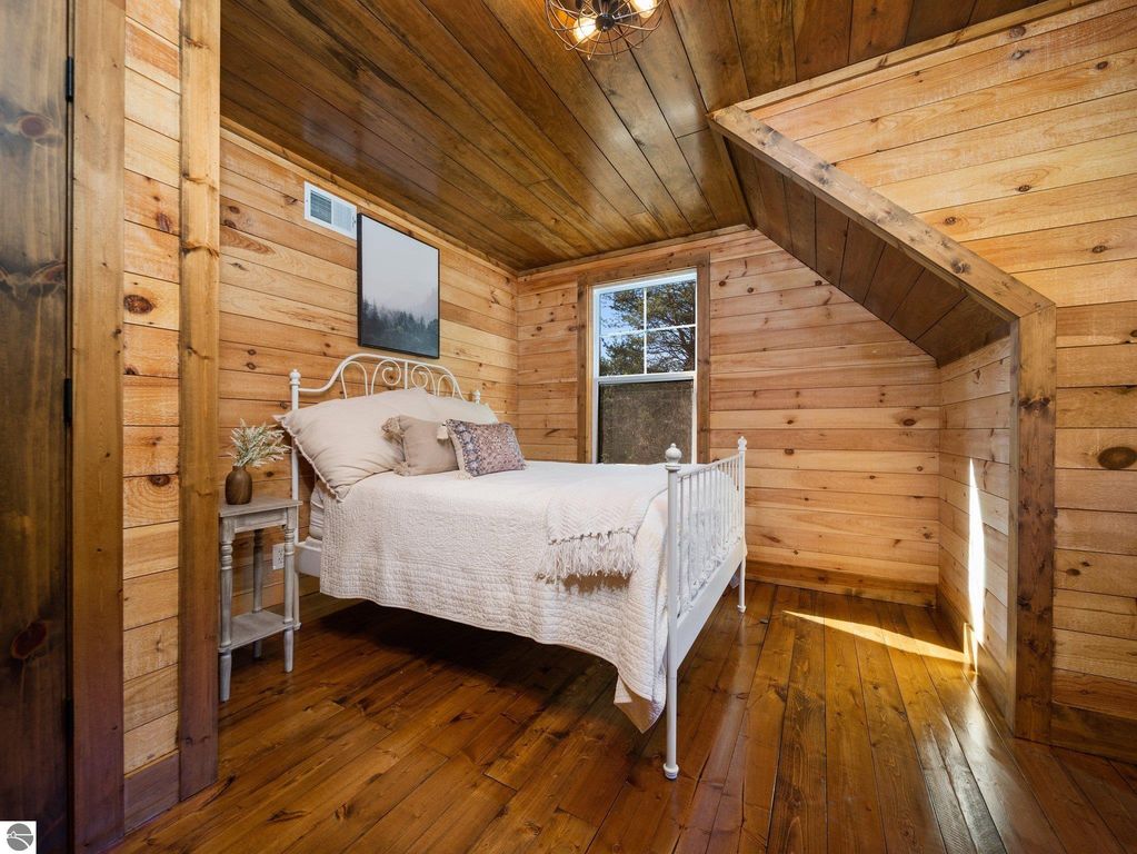 Cozy bedroom with wooden walls and ceiling, featuring a white metal bed, decorative pillows, and a small side table, showcasing natural light from the window and highlighting custom craftsmanship in a northern Michigan home.