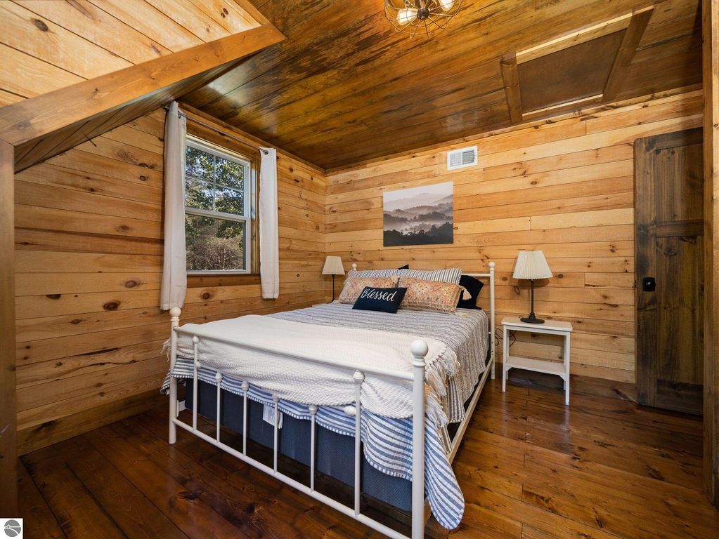 Cozy bedroom featuring a white metal bed with blue and striped bedding, wooden walls, and large windows, highlighting custom craftsmanship and rustic charm in a northern Michigan home.