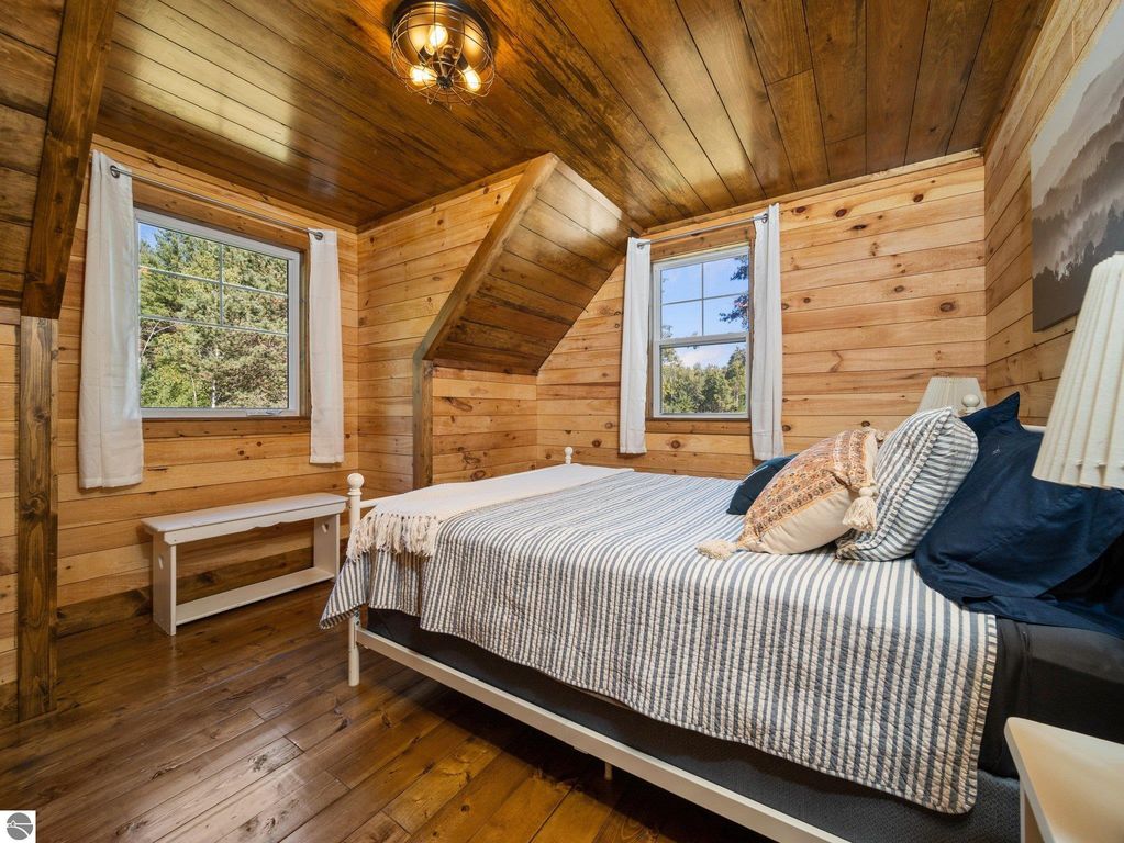 Cozy bedroom featuring a custom wood ceiling, natural wood paneling, a striped bedspread, and large windows offering views of northern Michigan's countryside.