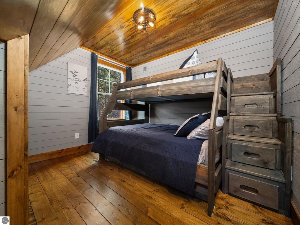 Cozy bedroom featuring a custom-built bunk bed with a lower full-size mattress, navy bedding, wooden accents, and a warm wood floor, showcasing northern Michigan craftsmanship and design.