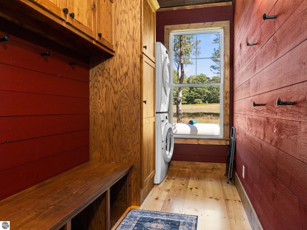 Mudroom with custom wood cabinetry, red shiplap walls, and large window showcasing northern Michigan countryside, featuring stacked washer and dryer, and wooden bench for storage.
