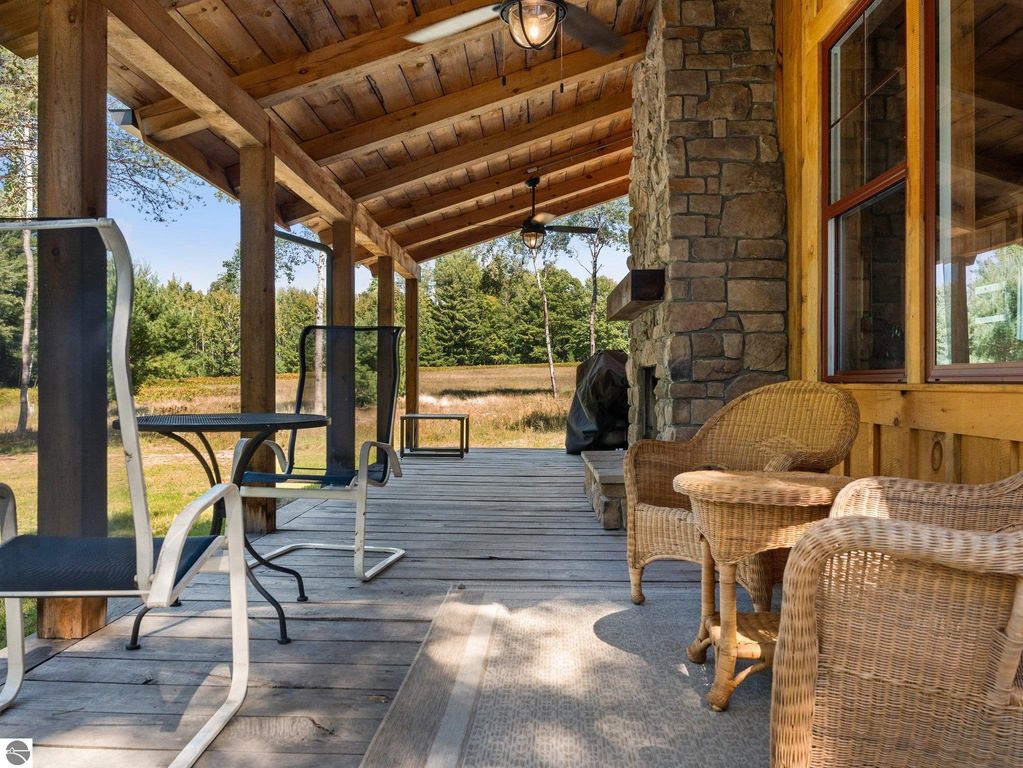Covered porch of a custom-built home in northern Michigan featuring wicker and metal chairs, a stone fireplace, and views of surrounding nature on a 10-acre property.