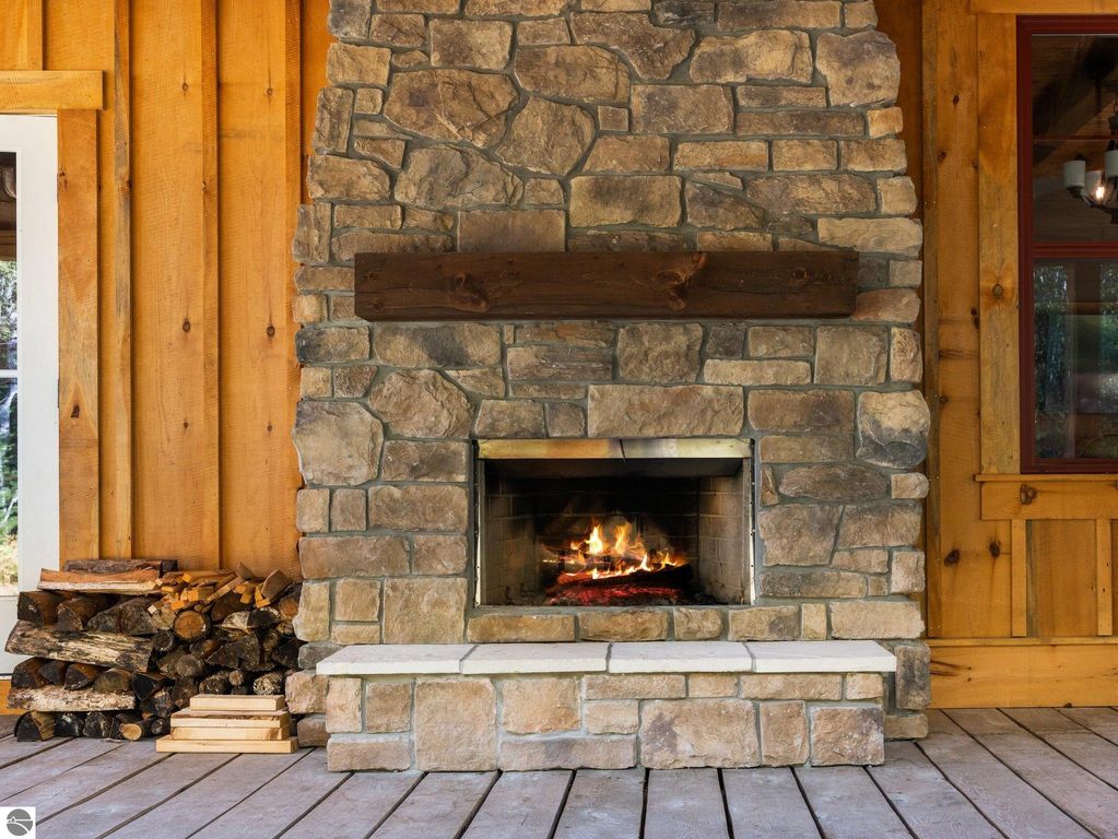 Cozy stone fireplace with wooden mantel, surrounded by rustic wood paneling and stacked firewood, enhancing the inviting atmosphere of the custom-built home in northern Michigan.