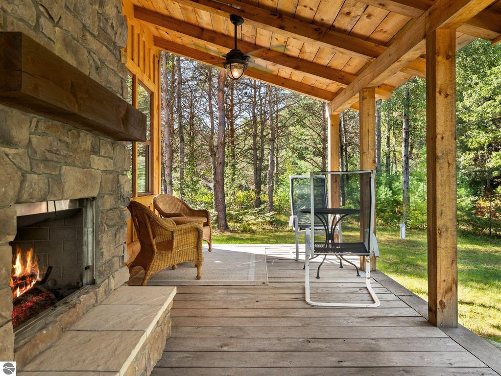 Covered porch with stone fireplace, wicker chair, and outdoor seating, surrounded by lush northern Michigan woods, showcasing peaceful countryside living at 5800 W Moorestown Road, Lake City, MI.