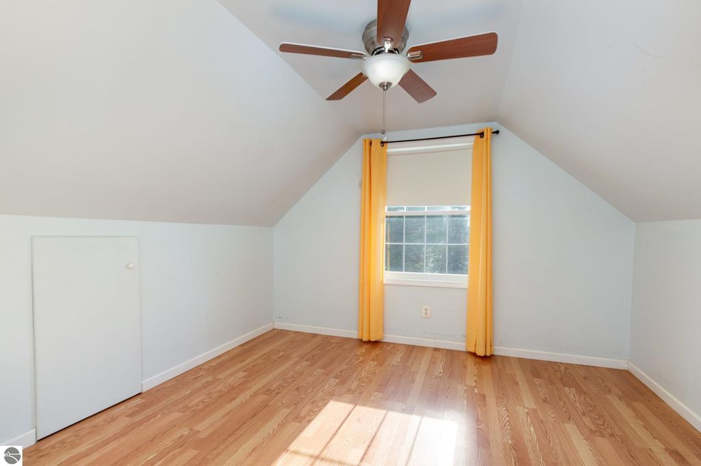 Spacious bonus room with sloped ceiling, hardwood floor, and large window featuring yellow curtains, ideal for guests or hobbies in Harrison, MI home listing.