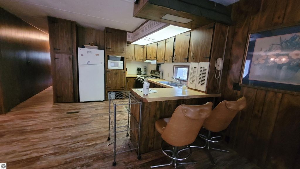 Kitchen interior with wooden cabinets, white appliances, and bar stools, showcasing the mobile home included in the Alden, MI real estate listing.