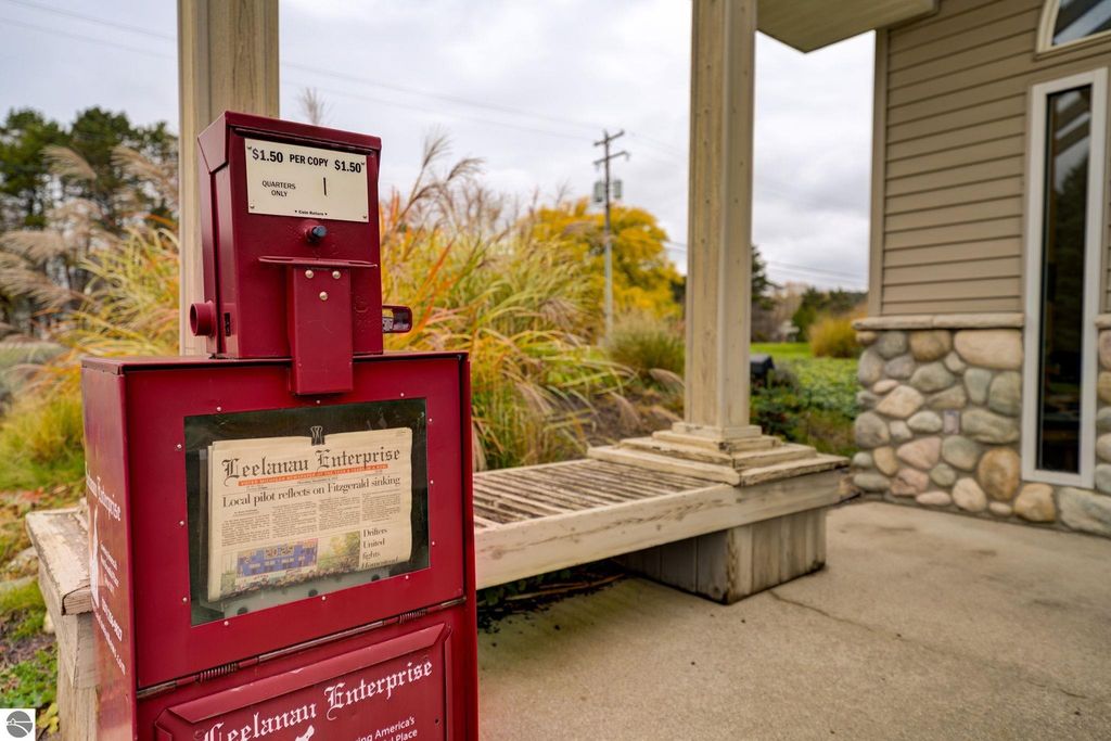Red newspaper vending machine displaying "Leelanau Enterprise" with local news, situated near commercial property at 7200 E Duck Lake Road, Lake Leelanau, MI.