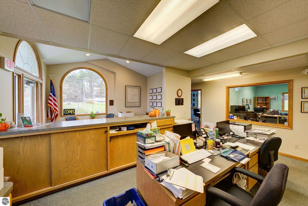 Reception area of a commercial building in Lake Leelanau, featuring a desk cluttered with paperwork, office supplies, and a "COPIES" sign, with an American flag and large windows visible in the background.