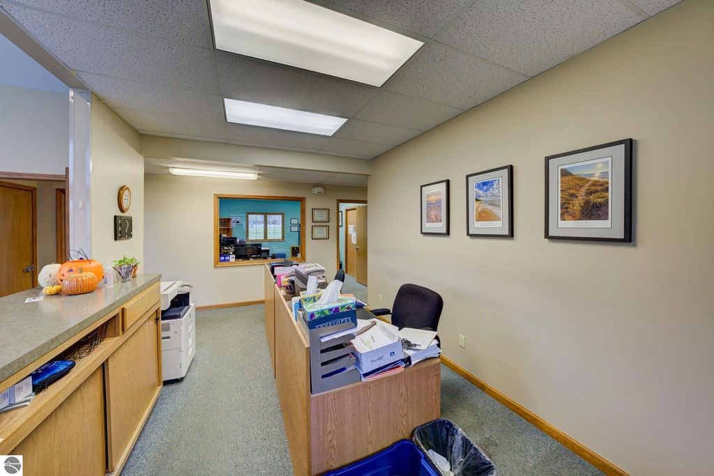 Reception area of a commercial office space with a desk, printer, and organized paperwork, featuring framed photographs on the wall and a view into an adjacent office, relevant to the commercial property listing at 7200 E Duck Lake Road, Lake Leelanau.