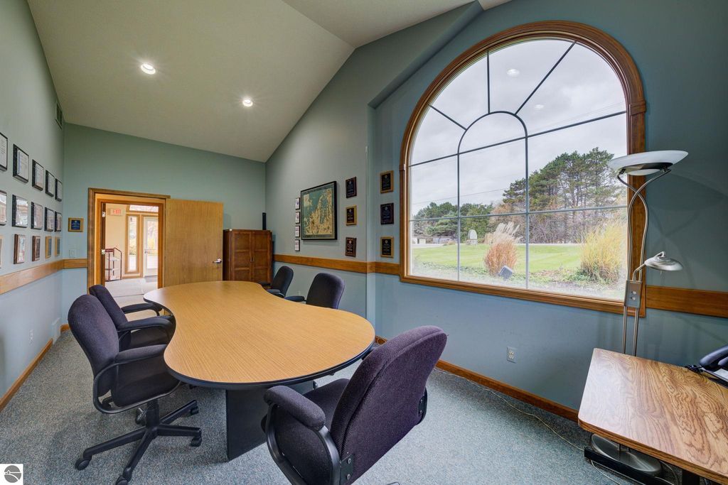 Conference room with a large oval table, black rolling chairs, wall-mounted plaques, and a large arched window overlooking green landscaping, highlighting the professional setting for the commercial property at 7200 E Duck Lake Road, Lake Leelanau, MI.