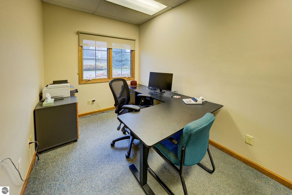 Office space interior featuring two desks, a computer monitor, and a printer, with a window providing natural light and a view of the surroundings, relevant to commercial property listing at 7200 E Duck Lake Road, Lake Leelanau, MI.