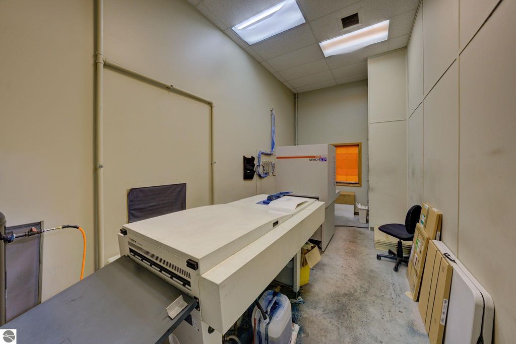 Interior view of commercial space featuring a large printing machine, workspace with office chair, and storage materials, illustrating potential for flexible build-out in Lake Leelanau property listing.