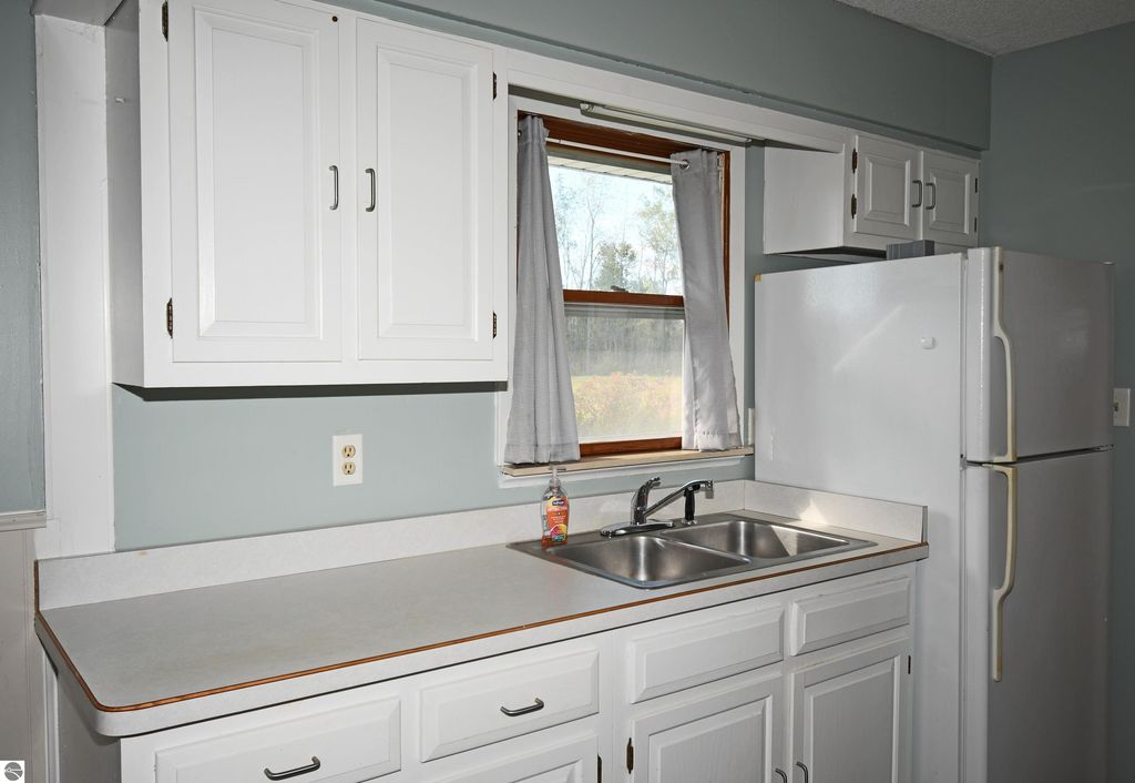Kitchen area featuring white cabinetry, double sink, and refrigerator, with a window offering views of the surrounding nature at the Tawas City ranch home for sale.