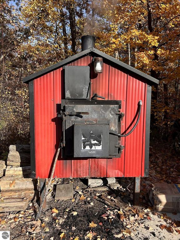 Red outdoor wood-burning stove with smoke rising, surrounded by autumn foliage, highlighting the cozy retreat atmosphere of the property at 5885 S Mt Tom Road, Rose City, MI.