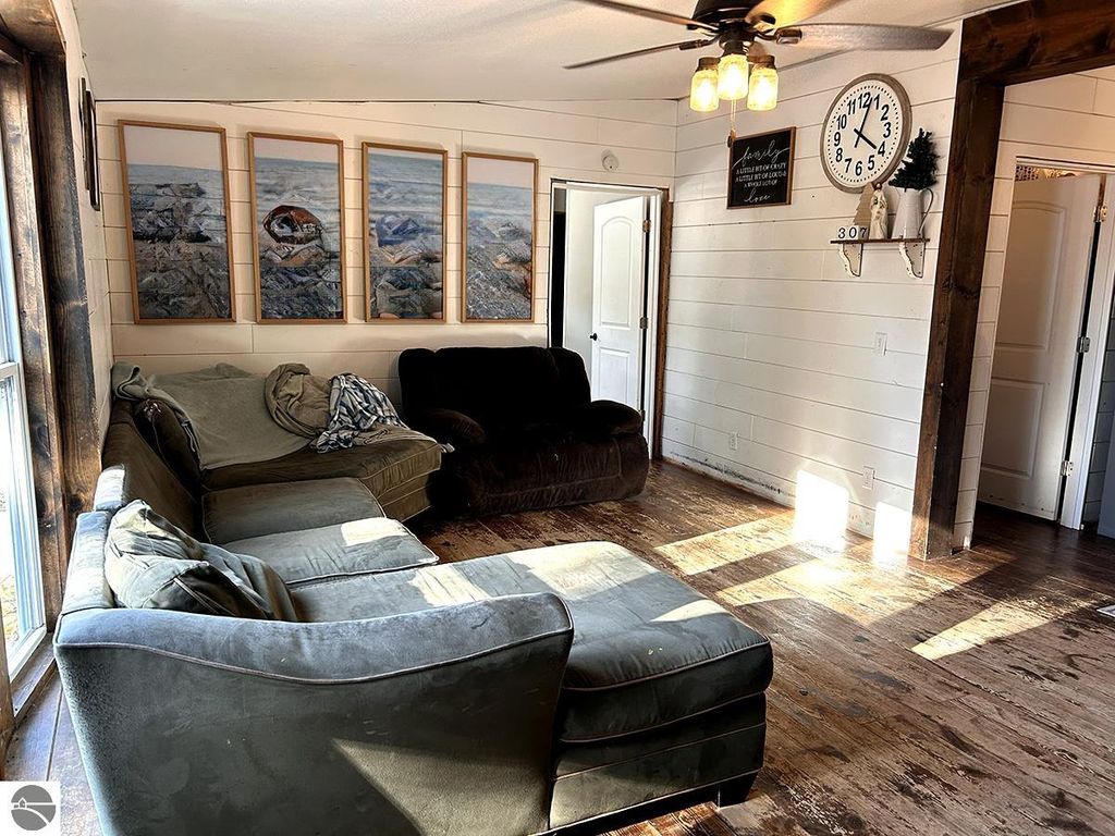 Cozy living room with green sectional sofa, framed nature artwork on walls, large clock, and natural light illuminating wooden floors, showcasing the inviting atmosphere of a 3-bedroom home for sale in Rose City, MI.
