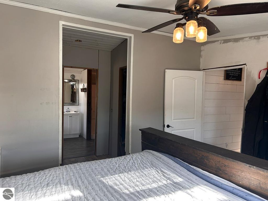 Interior view of a bedroom featuring a wooden bed frame, a ceiling fan with exposed bulbs, and an open doorway leading to a bathroom with a mirror and vanity, showcasing potential for a cozy up north retreat in Rose City, MI.