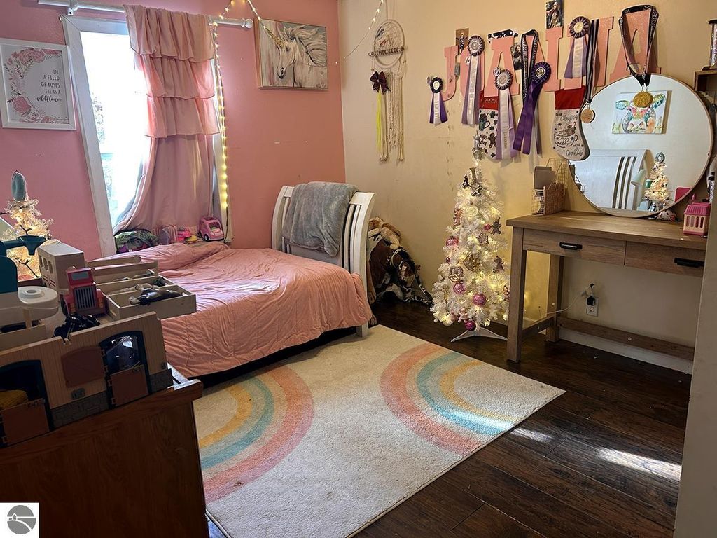 Cozy bedroom with pink walls, bed with pink bedding, small white Christmas tree, and decorated wall showcasing ribbons and awards.