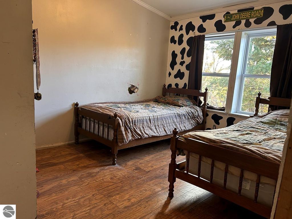 Cozy bedroom with two wooden beds, quilted bedding, and cow-patterned wall decor, featuring a large window overlooking trees, ideal for a peaceful retreat in Rose City, MI.