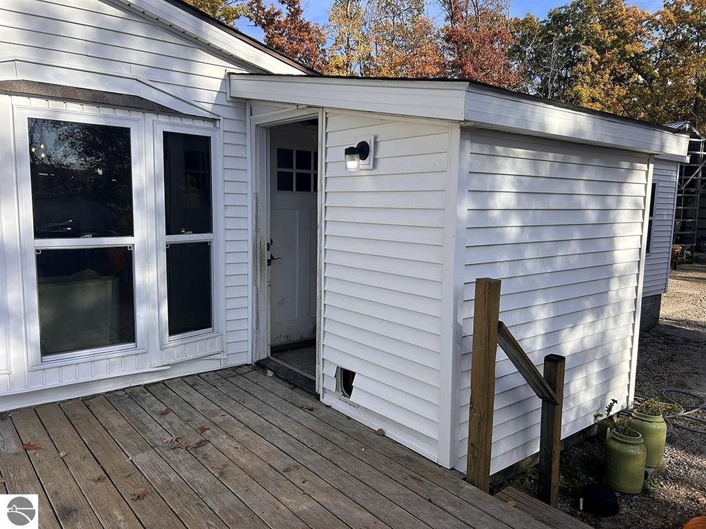 Exterior view of a white house entrance with large windows, wooden deck, and a side door, surrounded by trees showcasing autumn foliage, emphasizing the cozy retreat aspect of the property for sale at 5885 S Mt Tom Road, Rose City, MI.