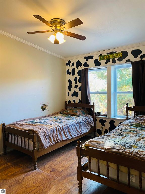Cozy bedroom with two wooden beds, cow-patterned wall decor, and a ceiling fan, featuring large windows with dark curtains, highlighting a rustic Up North retreat ambiance.