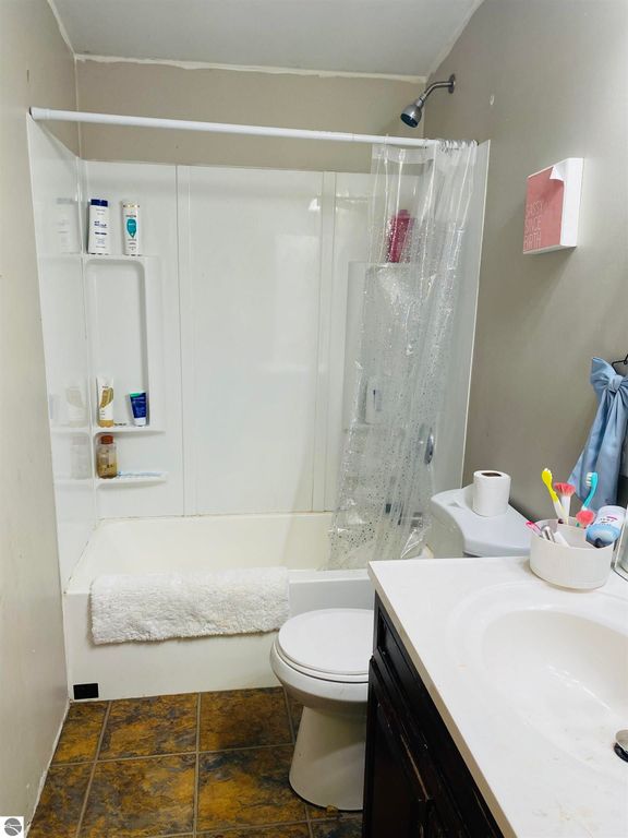Bathroom interior featuring a shower-tub combination, toilet, and sink with toiletries and cleaning supplies, showcasing a cozy, functional space in a 3-bedroom home for sale in Rose City, MI.