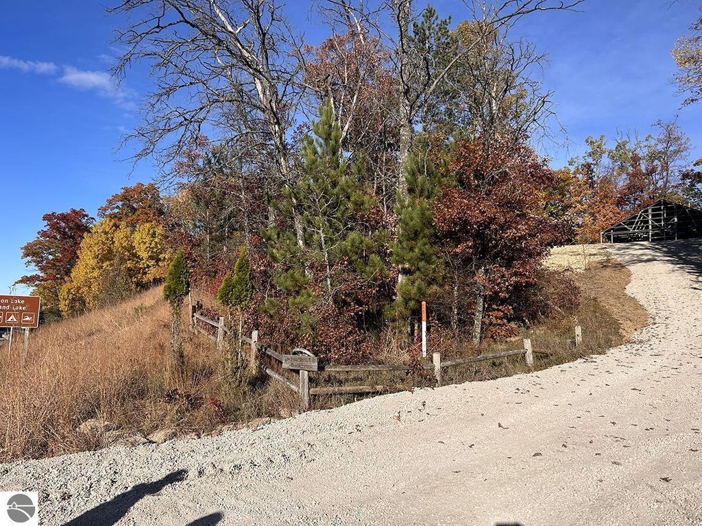 Scenic view of a gravel road leading to a wooded area near Island Lake and Loon Lake, with autumn foliage, wooden fencing, and a directional sign indicating nearby lakes, emphasizing outdoor recreational opportunities in Rose City, MI.
