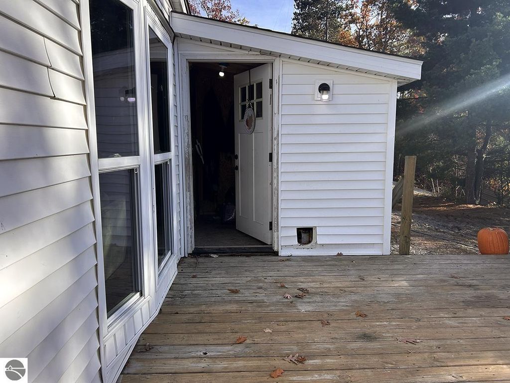 Entrance view of a cozy home at 5885 S Mt Tom Road, Rose City, MI, featuring a white exterior, wooden deck, and inviting doorway, surrounded by autumn foliage and a pumpkin on the porch.