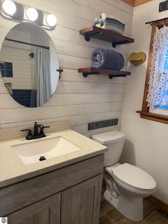 Chalet-style bathroom with modern fixtures, oval mirror, wooden shelves holding towels, and a sign reading "PRIMITIVES," showcasing a cozy and updated aesthetic in a home near Torch Lake.