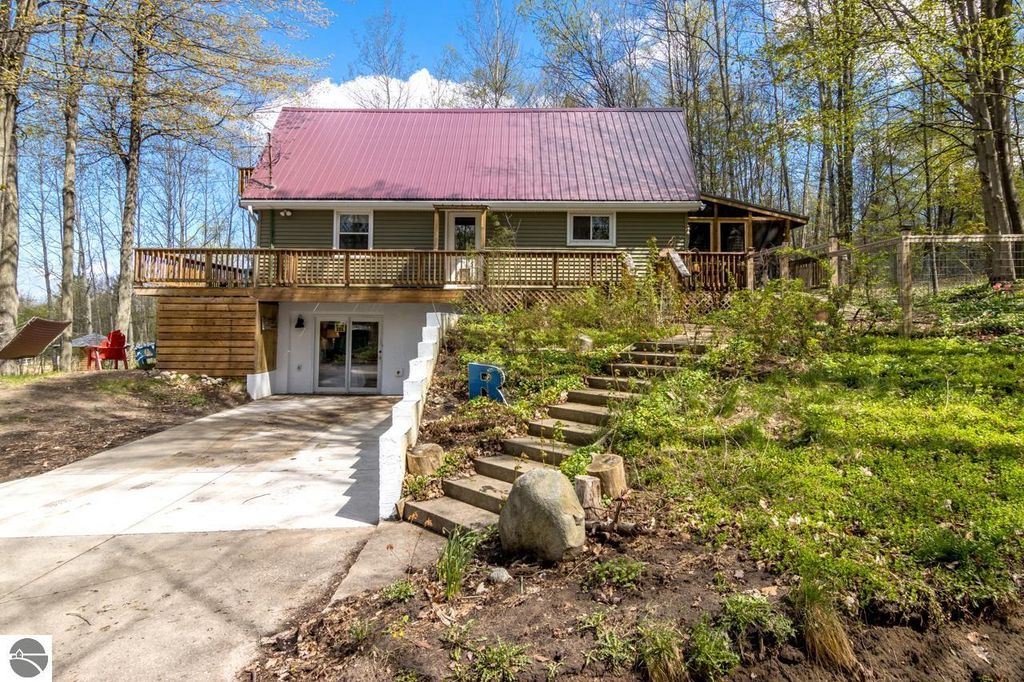 Chalet style home on 5 acres in Alden, MI, featuring a red metal roof, wooden deck, and natural landscaping, ideal for year-round living near Torch Lake.