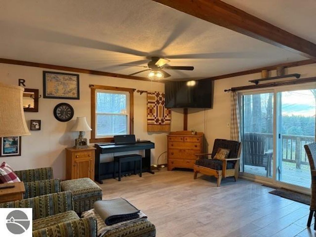 Chalet-style living room featuring a piano, comfortable seating, wall decor, and large windows with views of the natural surroundings, emphasizing the cozy ambiance of the property at 9535 Serenity Trail, Alden, MI.