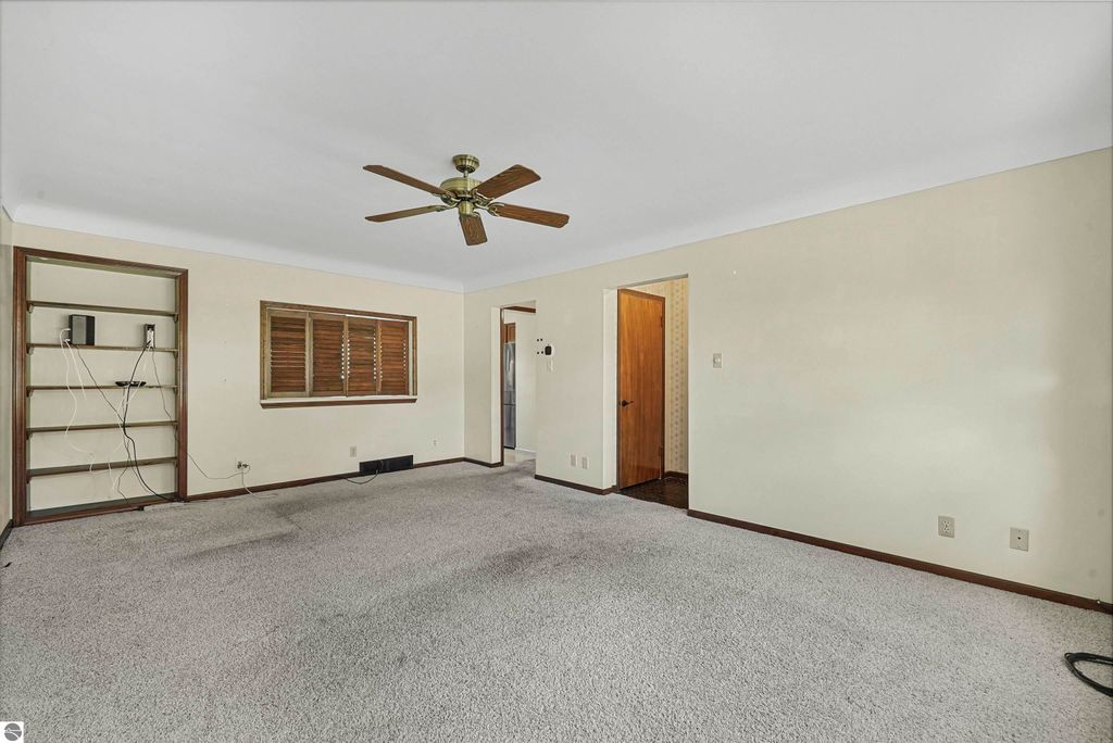 Living room of a ranch-style home at 315 N Simons Street, Cadillac, MI, featuring beige walls, carpeted flooring, a ceiling fan, and a window with wooden shutters, showcasing a spacious layout suitable for family living.