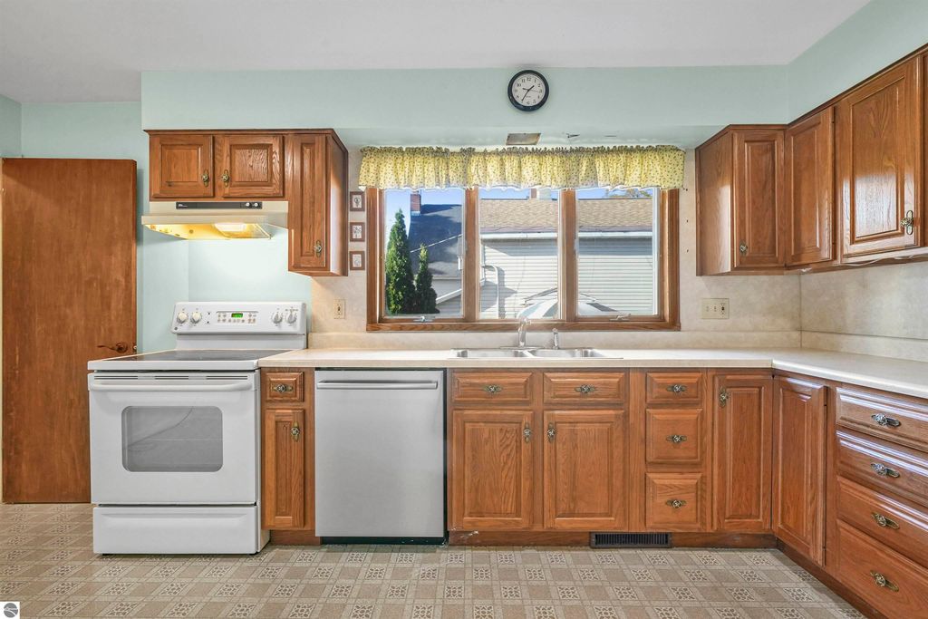 Kitchen interior featuring wooden cabinets, white appliances, and a window with a yellow curtain, showcasing a clean and functional space in a ranch home for sale at 315 N Simons Street, Cadillac, MI.