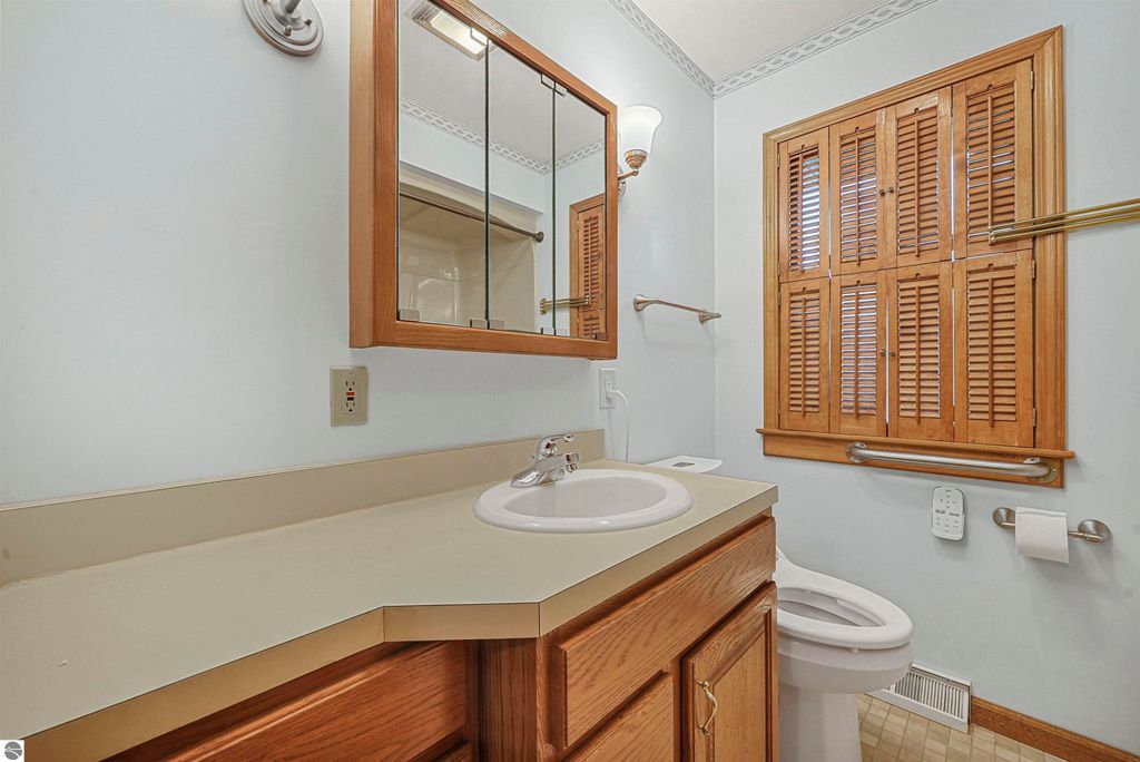 Bathroom with wooden cabinetry, sink, and mirror, featuring a window with wooden shutters and light blue walls, showcasing a clean and well-maintained space in a Cadillac, MI home listing.