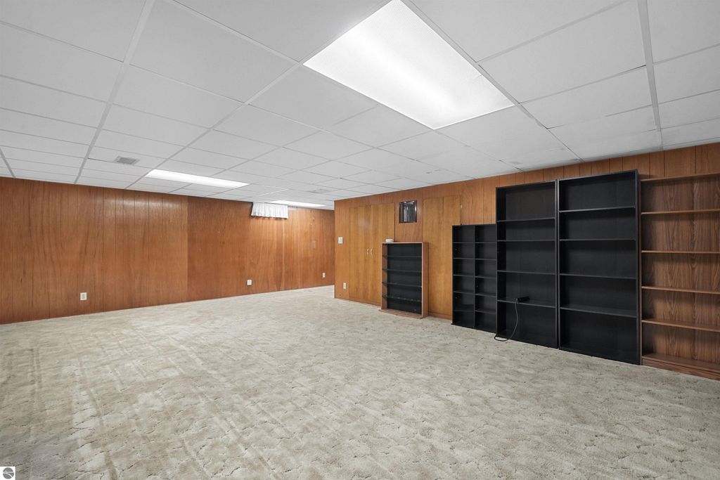 Finished basement with beige carpet, wooden panel walls, and black shelving units, showcasing additional living space in a 3-bedroom, 2-bath ranch home in Cadillac, MI.
