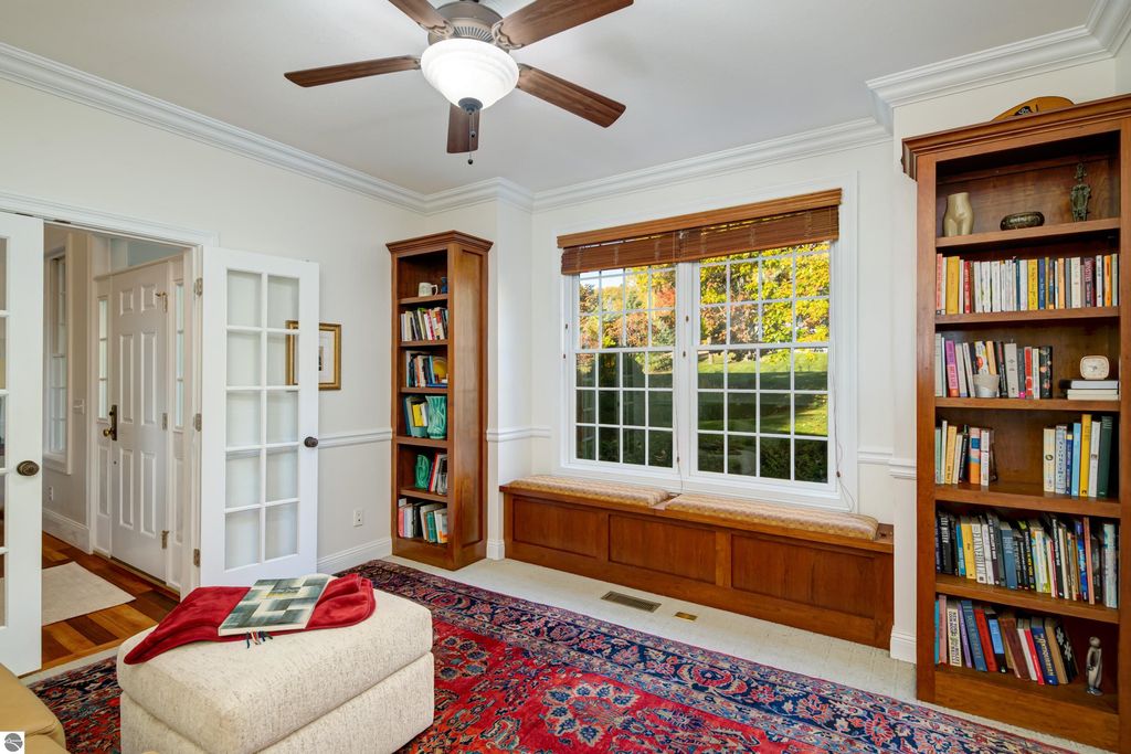 Cozy reading nook with built-in bookshelf, plush ottoman, and large windows showcasing landscaped yard, in a custom-designed home at 6436 Peregrin Court, Traverse City.