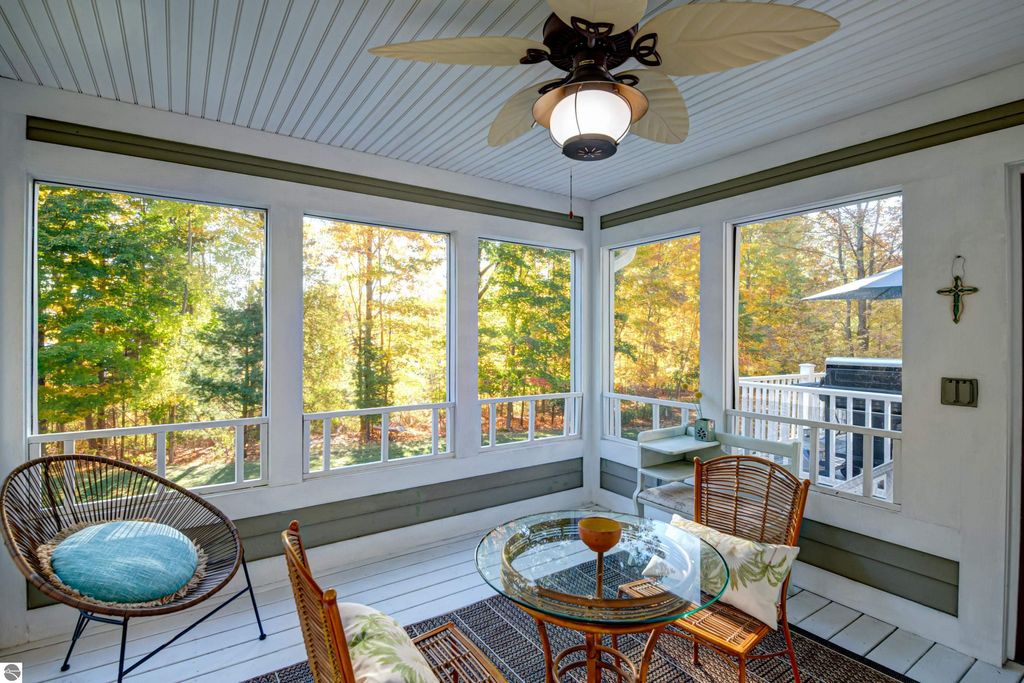 Sunlit three-season sunroom with large windows showcasing vibrant fall foliage, featuring a round glass table, wicker chairs, and a ceiling fan, enhancing the inviting atmosphere of the Traverse City property at 6436 Peregrin Court.