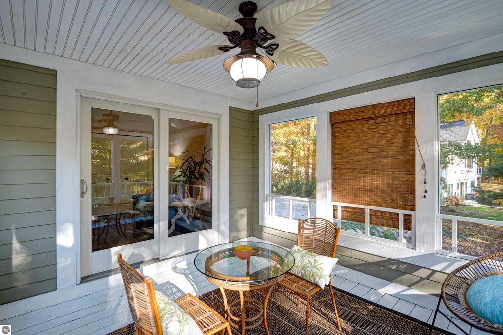 Sunroom with ceiling fan, glass table, and wicker chairs, showcasing bright natural light and views of a landscaped yard, part of the property at 6436 Peregrin Court, Traverse City, MI.
