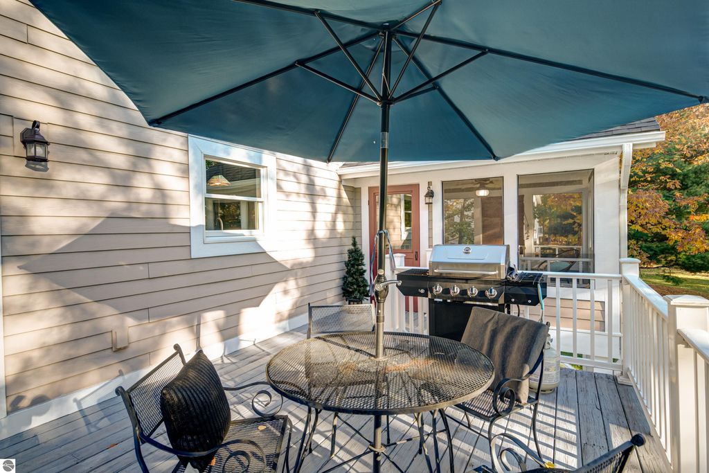Outdoor patio with a table and chairs under a blue umbrella, featuring a grill, adjacent to a home with a landscaped yard, highlighting the inviting outdoor living space at 6436 Peregrin Court, Traverse City, MI.