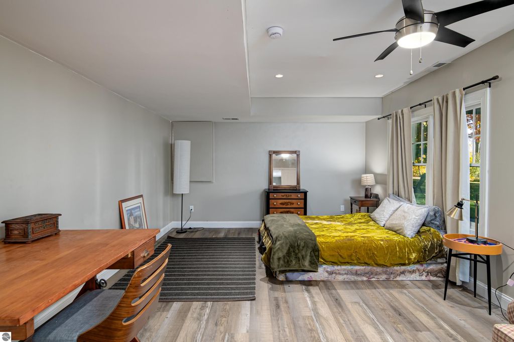 Spacious bedroom featuring a cozy bed with a green throw, wooden desk, and natural light from large windows, showcasing modern comfort in Traverse City home for sale at 6436 Peregrin Court.