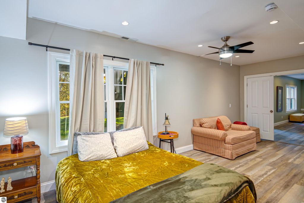 Cozy bedroom with gold bedding, large windows, and a comfortable seating area featuring a plaid sofa, showcasing modern design elements in a home for sale at 6436 Peregrin Court, Traverse City, MI.