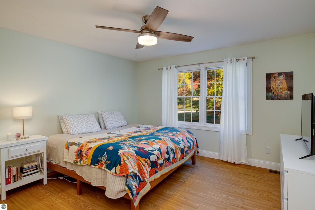 Brightly lit bedroom featuring a large bed with a colorful floral quilt, bedside table with lamp and books, large window with white curtains, and a modern ceiling fan, showcasing the cozy ambiance of the property at 6436 Peregrin Court in Traverse City, MI.